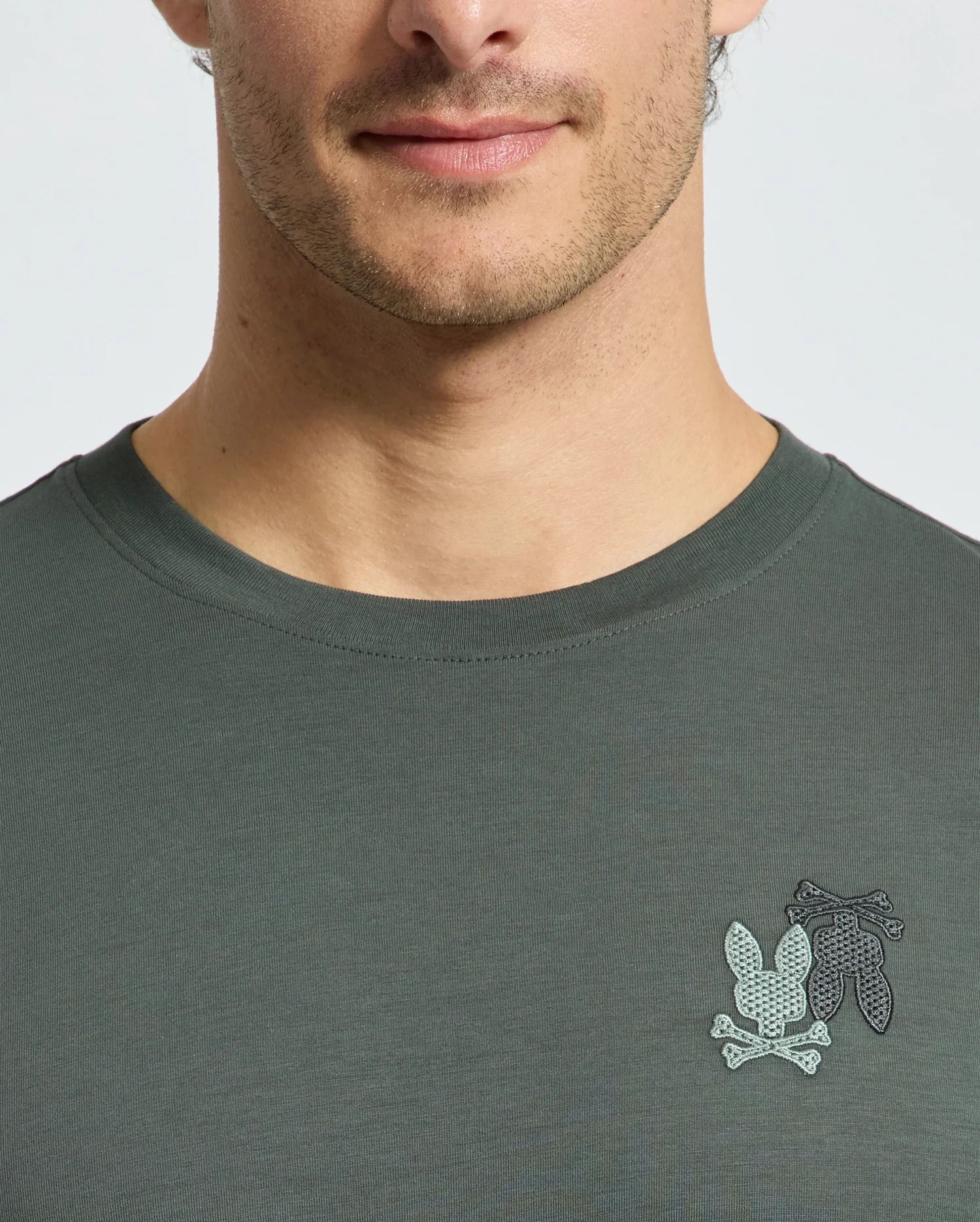 The image shows a person in a dark green Psycho Bunny MENS PIERCE LONG SLEEVE BACK GRAPHIC TEE, made of Pima cotton, with three cacti and two crossed bones on the left chest. They are captured from the shoulders up with a neutral expression.