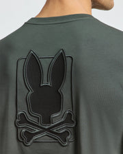 Someone is wearing the Psycho Bunny MENS PIERCE LONG SLEEVE BACK GRAPHIC TEE - B6T178E200, a dark green shirt crafted from soft Pima cotton. It features a shiny black embroidered bunny silhouette above crossed bones on the back, contrasting beautifully with the matte fabric.