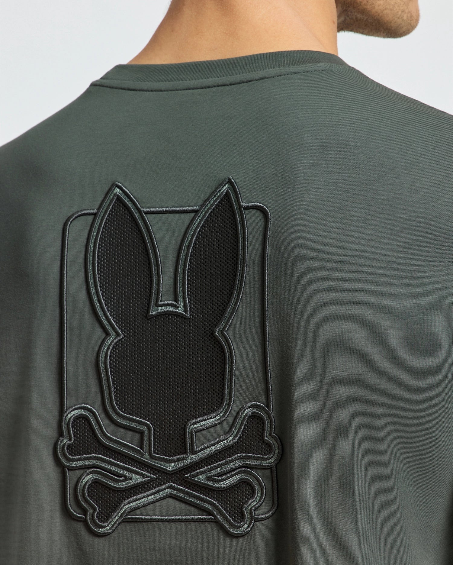 Someone is wearing the Psycho Bunny MENS PIERCE LONG SLEEVE BACK GRAPHIC TEE - B6T178E200, a dark green shirt crafted from soft Pima cotton. It features a shiny black embroidered bunny silhouette above crossed bones on the back, contrasting beautifully with the matte fabric.