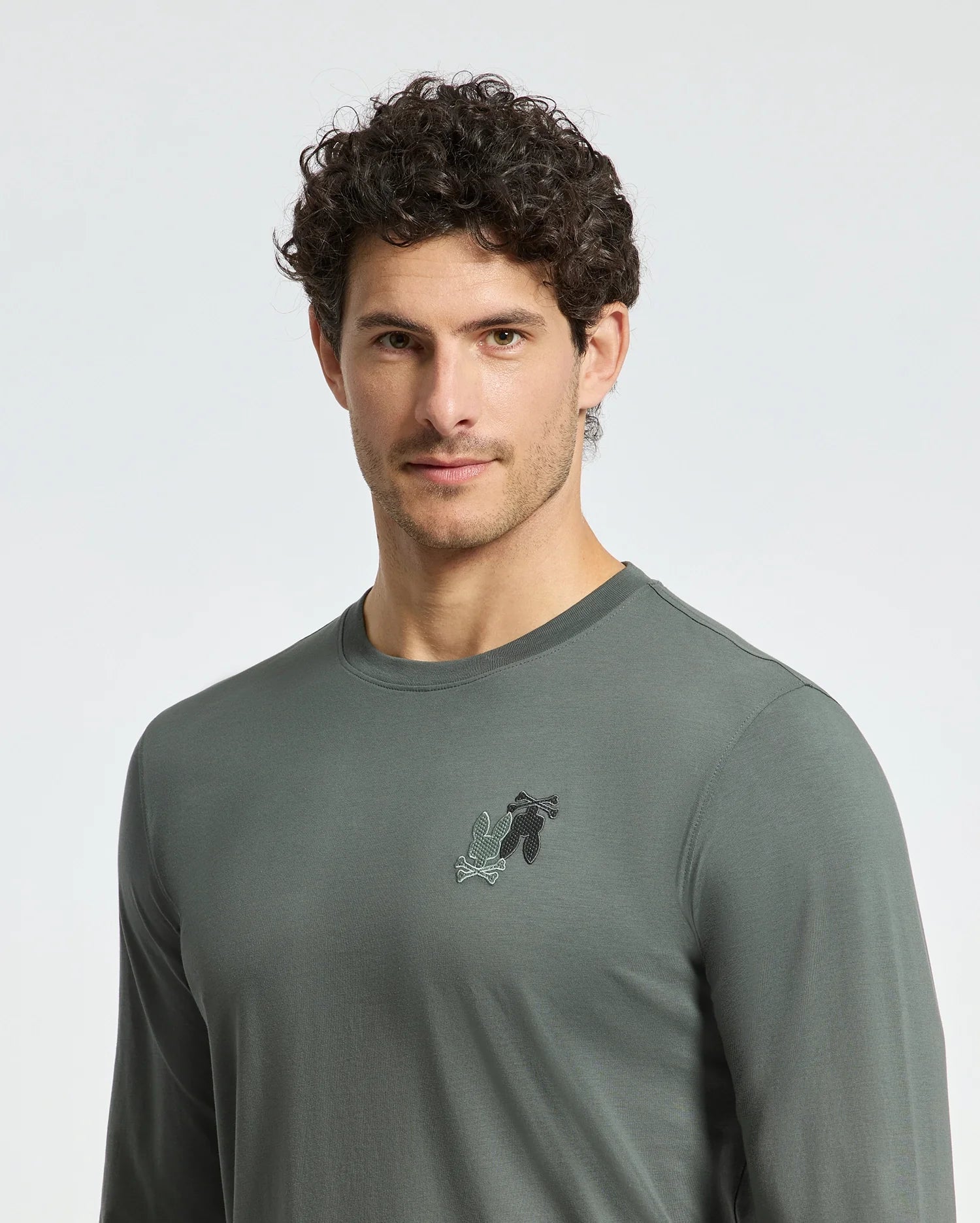 A man with curly hair wears a Psycho Bunny MENS PIERCE LONG SLEEVE BACK GRAPHIC TEE - B6T178E200, crafted from comfortable Pima cotton, featuring an embroidered design on the left chest. He stands against a light background, embodying casual elegance.
