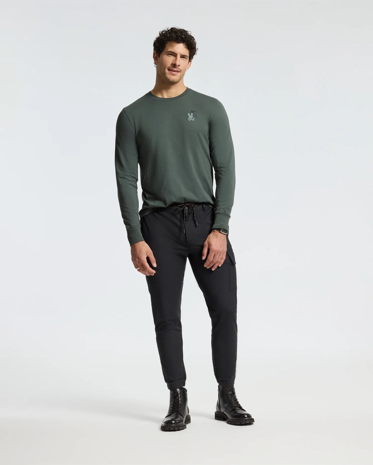 A person in a Psycho Bunny MENS PIERCE LONG SLEEVE BACK GRAPHIC TEE and black jogger pants stands against a plain background, featuring short curly hair and black boots.
