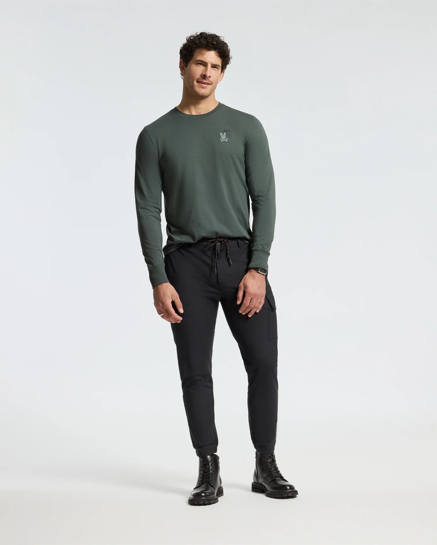 A person in a Psycho Bunny MENS PIERCE LONG SLEEVE BACK GRAPHIC TEE and black jogger pants stands against a plain background, featuring short curly hair and black boots.