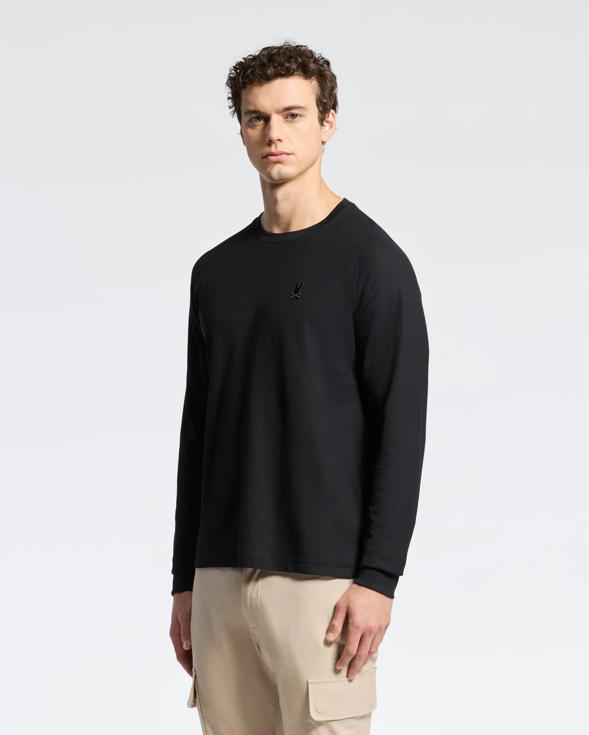 A man in the Psycho Bunny MENS TAYLOR PIQUE LONG SLEEVE TEE in sleek black and beige cargo pants stands against a white background, highlighted by an embroidered Bunny for added charm.