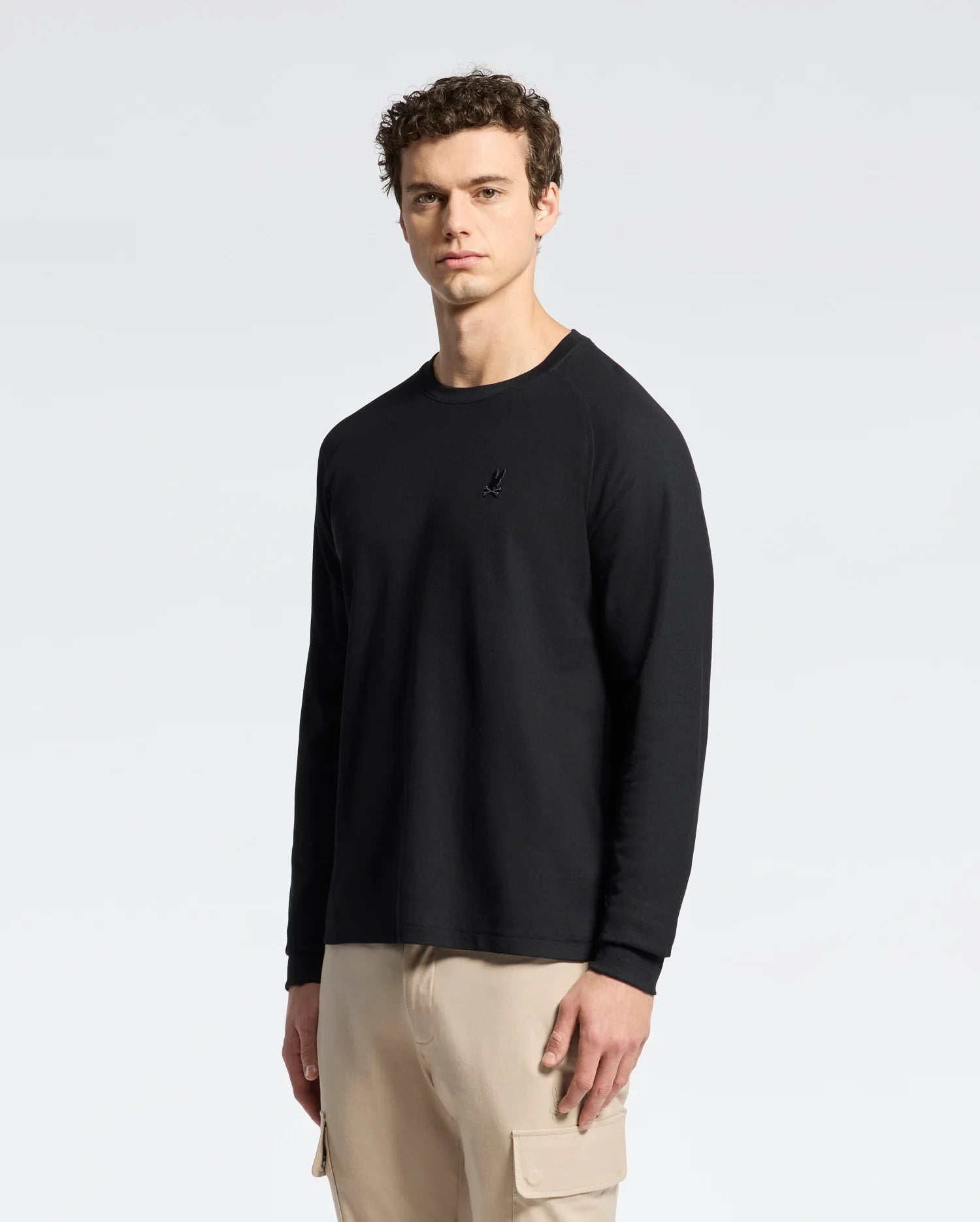 A man in the Psycho Bunny MENS TAYLOR PIQUE LONG SLEEVE TEE in sleek black and beige cargo pants stands against a white background, highlighted by an embroidered Bunny for added charm.