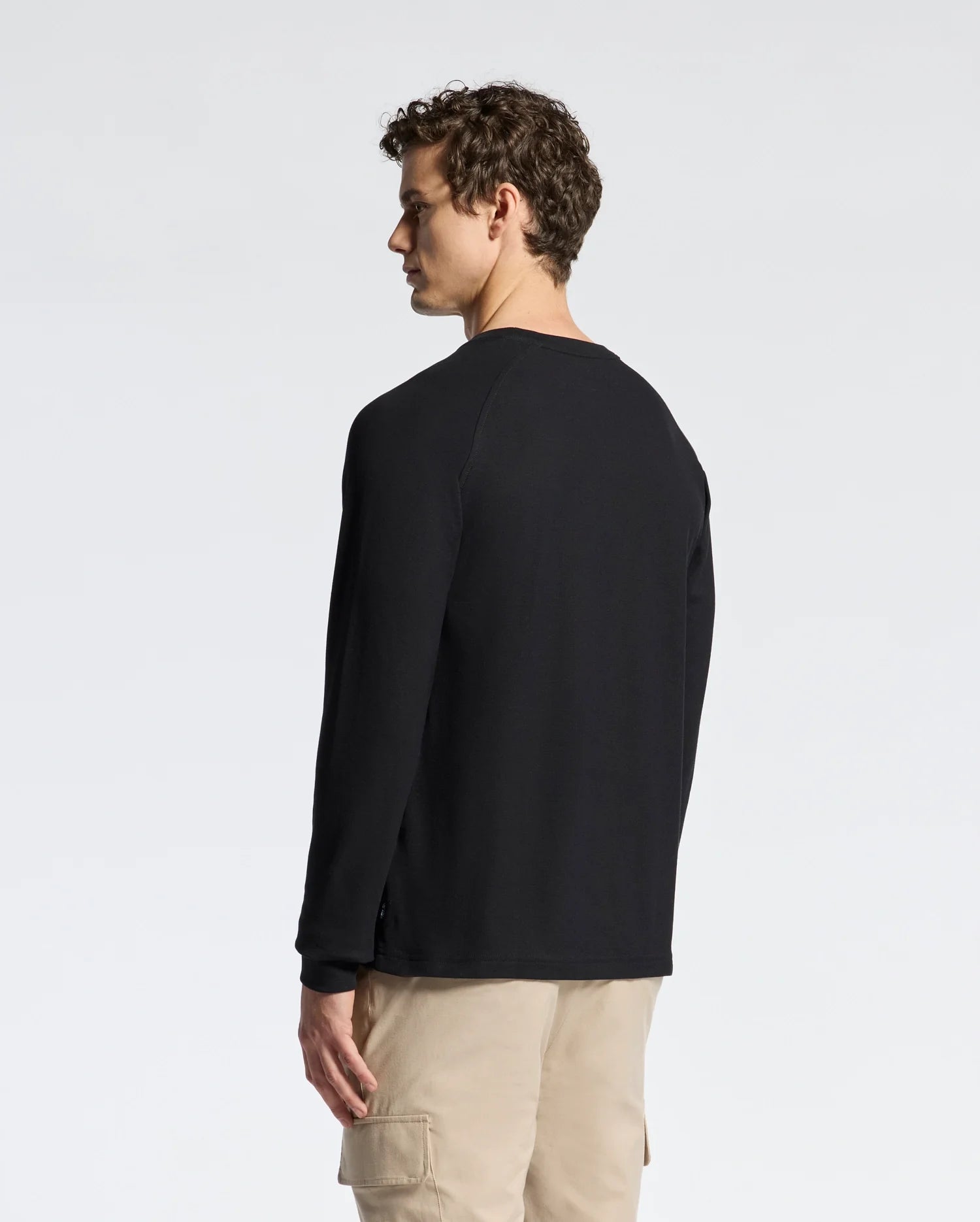 A person with curly hair faces away from the camera in a black Psycho Bunny long-sleeve Taylor Pique tee, paired with beige cargo pants against a light gray backdrop.