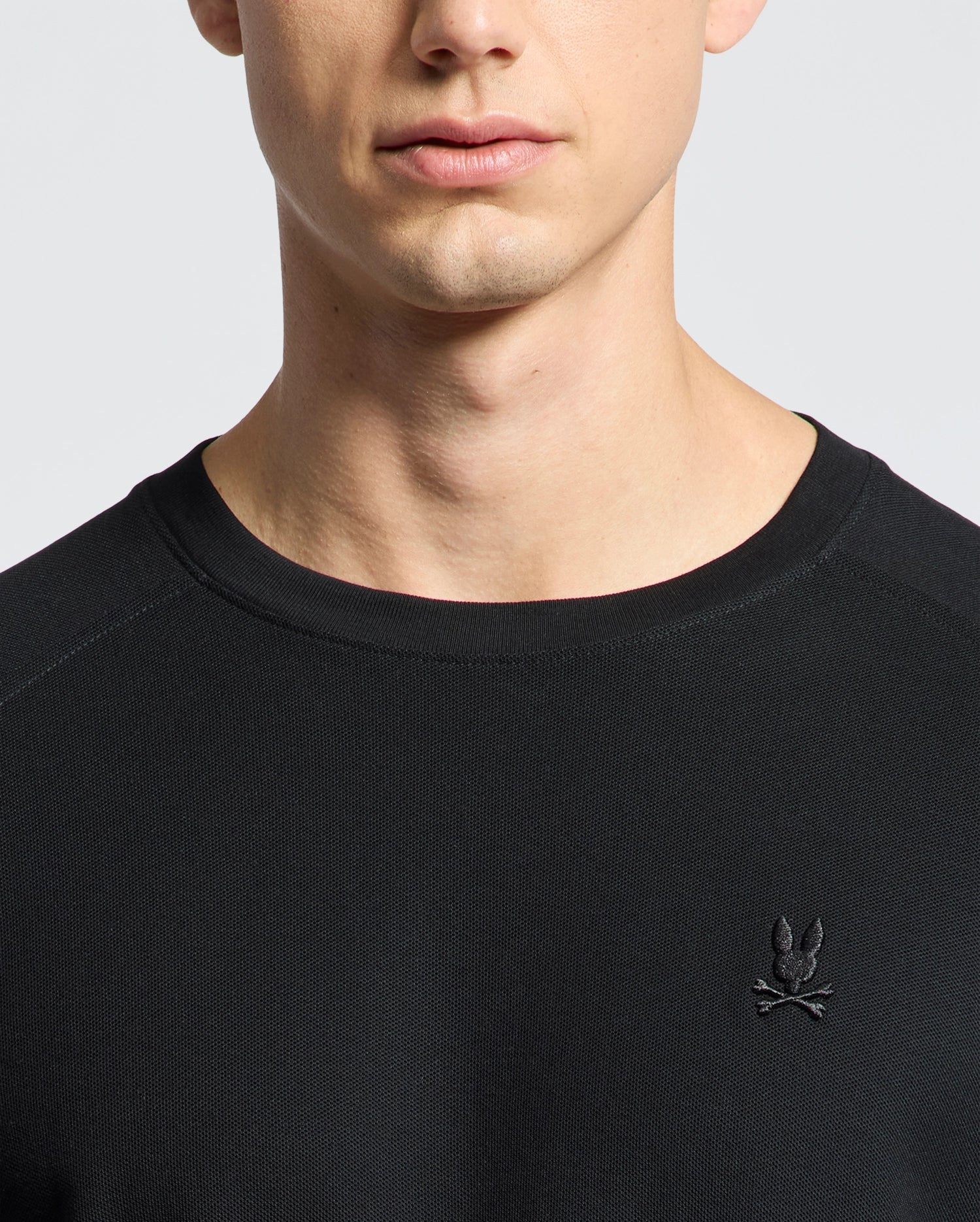 A person wears a Psycho Bunny black Pima cotton piqué long-sleeve tee, featuring an embroidered Bunny with crossed bones on the chest. The background is plain and light-colored, highlighting the upper torso and neck area.