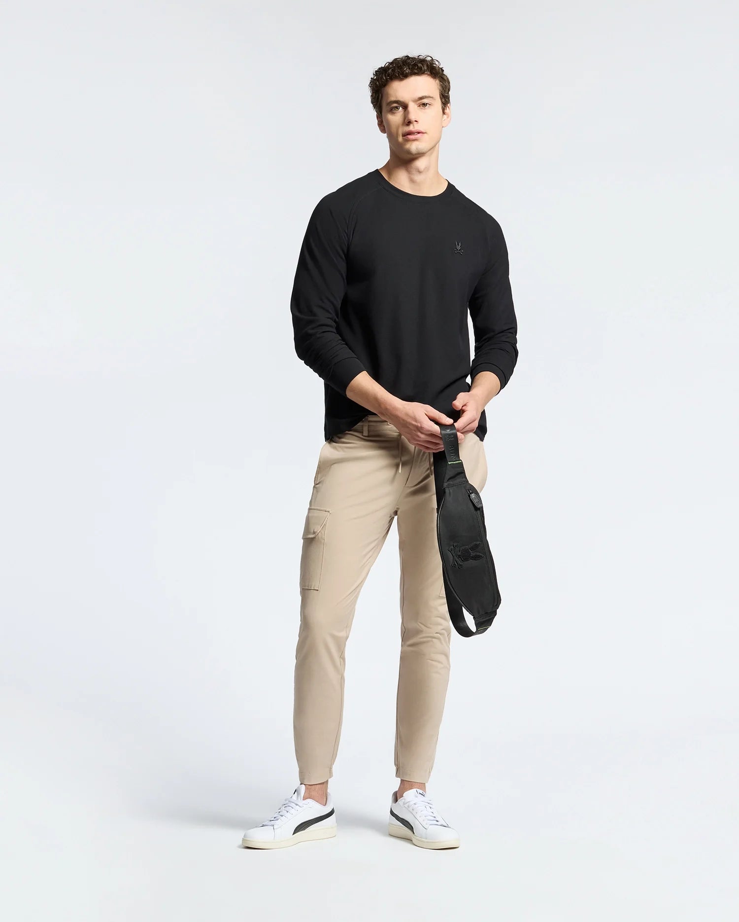 A man stands against a simple background wearing a black Psycho Bunny Men's Taylor Piqué Long Sleeve Tee, beige cargo pants, and white sneakers. He holds an embroidered Bunny crossbody bag in his left hand and looks towards the camera.