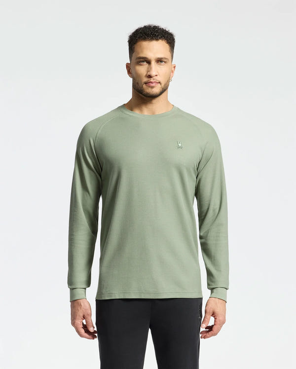 A person with short curly hair stands against a plain background, wearing Psycho Bunny's men's Taylor piqué long-sleeve tee and black pants. The light green shirt features a small embroidered logo on the left chest.