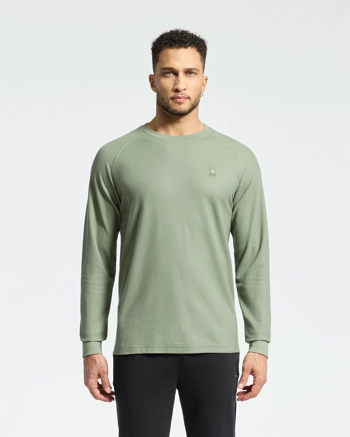 A person with short curly hair stands against a plain background, wearing Psycho Bunny's men's Taylor piqué long-sleeve tee and black pants. The light green shirt features a small embroidered logo on the left chest.