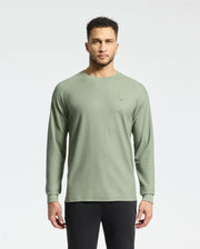 A person with short curly hair stands against a plain background, wearing Psycho Bunny's men's Taylor piqué long-sleeve tee and black pants. The light green shirt features a small embroidered logo on the left chest.