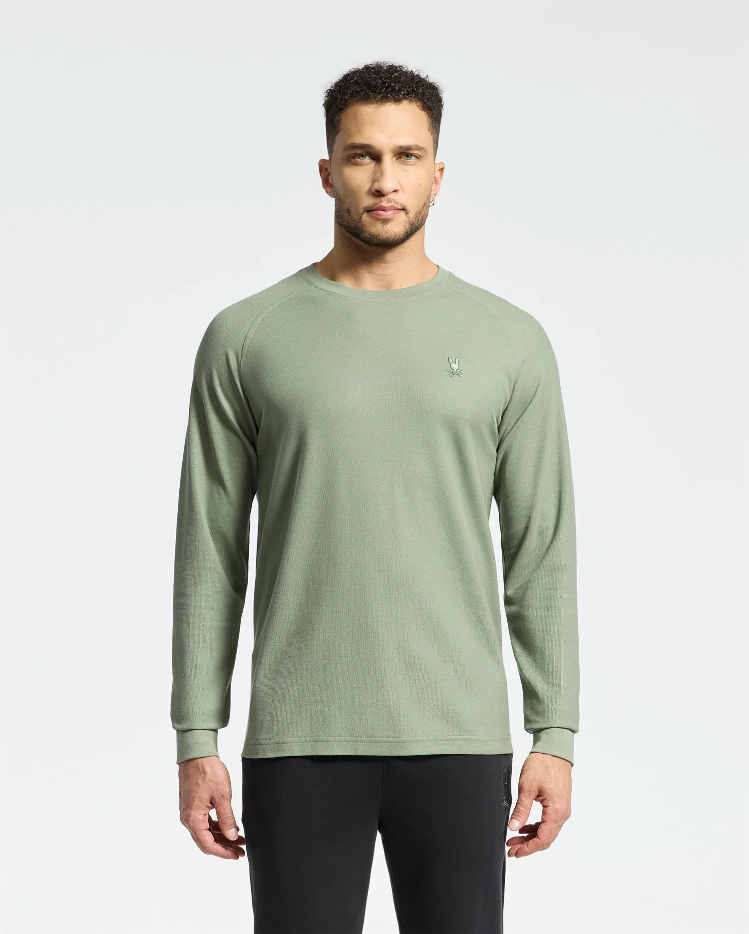 A person with short curly hair stands against a plain background, wearing Psycho Bunny's men's Taylor piqué long-sleeve tee and black pants. The light green shirt features a small embroidered logo on the left chest.