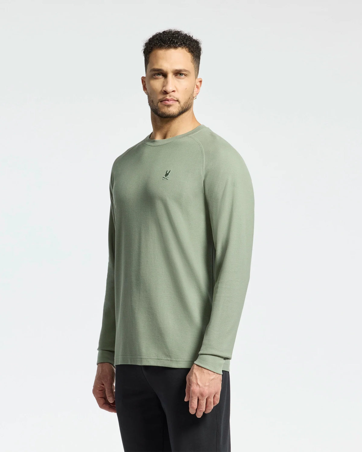 A man in a Psycho Bunny MENS TAYLOR PIQUE LONG SLEEVE TEE - B6T580F200, made from light olive green Pima cotton with a small chest logo, stands against a white background wearing black pants and displaying a neutral expression.
