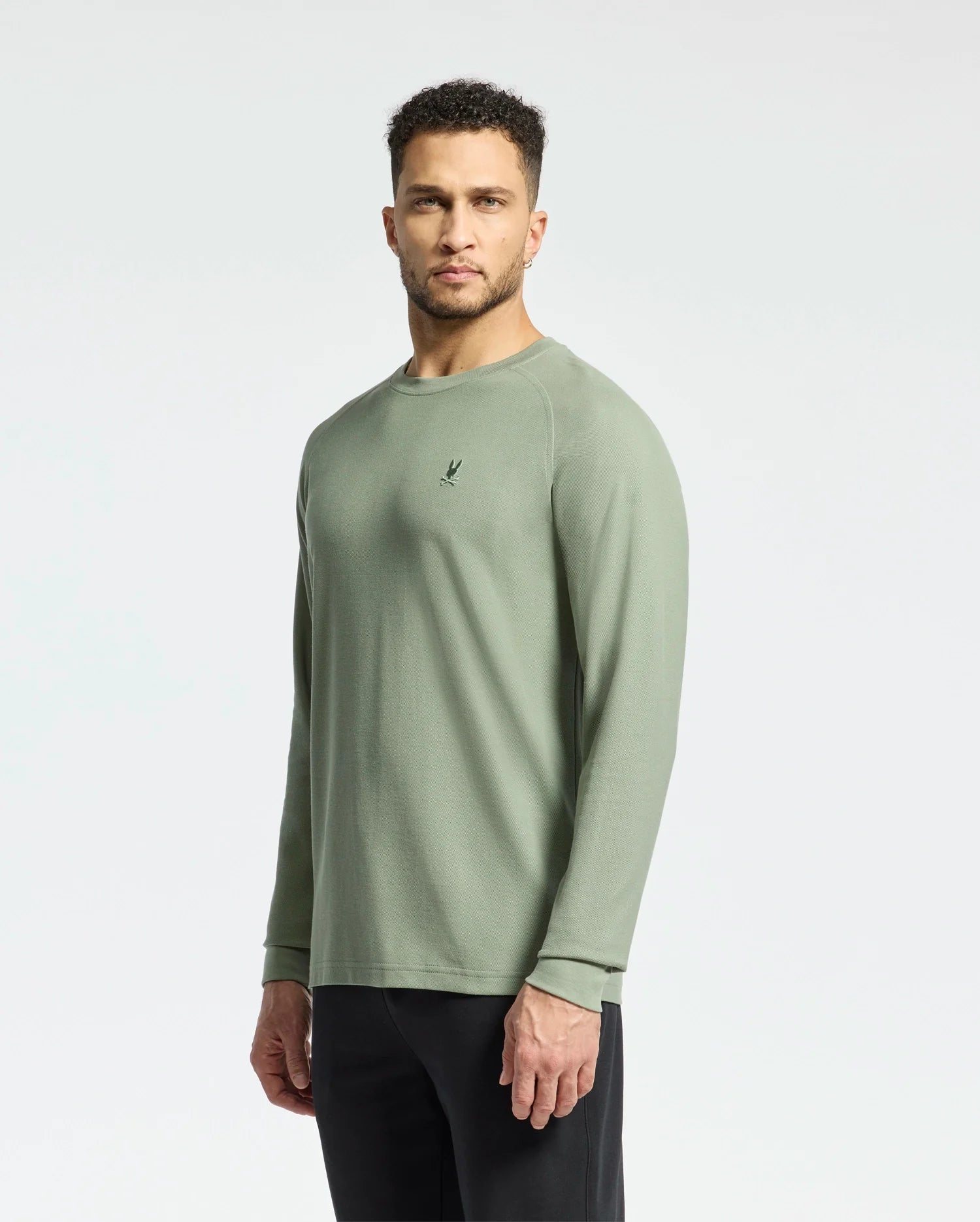 A man in a Psycho Bunny MENS TAYLOR PIQUE LONG SLEEVE TEE - B6T580F200, made from light olive green Pima cotton with a small chest logo, stands against a white background wearing black pants and displaying a neutral expression.