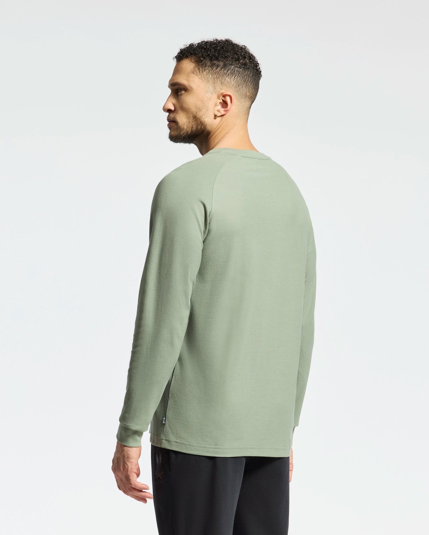 A person with short curly hair stands against a plain white background, wearing the Psycho Bunny Men's Taylor Pique Long Sleeve Tee (B6T580F200) in light green and black pants. They are facing slightly to the side, looking away.