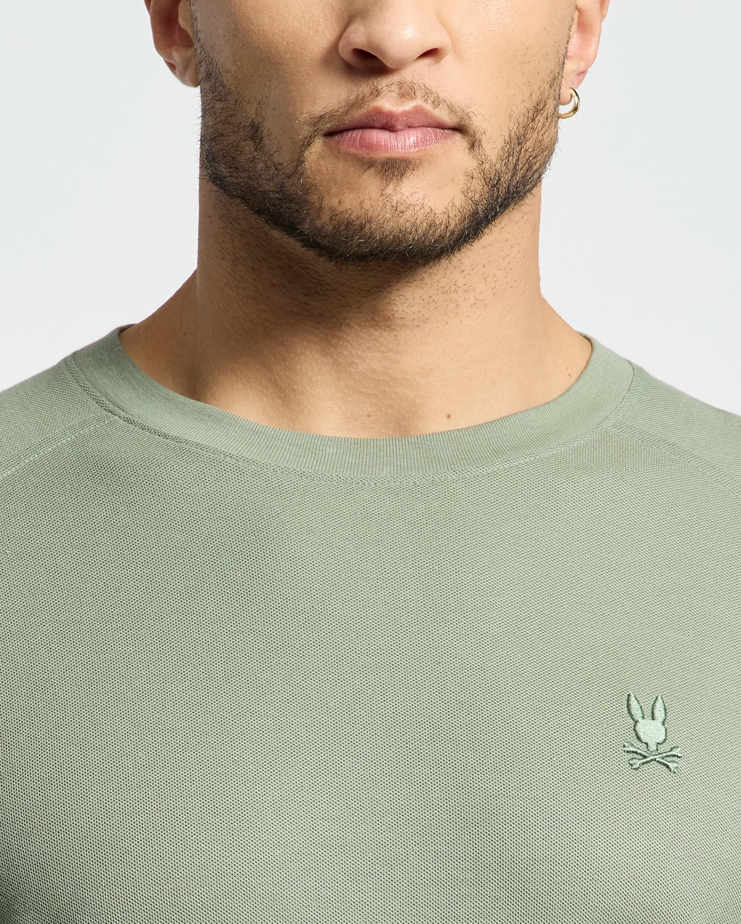 A man in a sage green Psycho Bunny MENS TAYLOR PIQUE LONG SLEEVE TEE with a rabbit and bones logo on the left. He has a beard and wears a small hoop earring. The image focuses on his lower face and upper torso.
