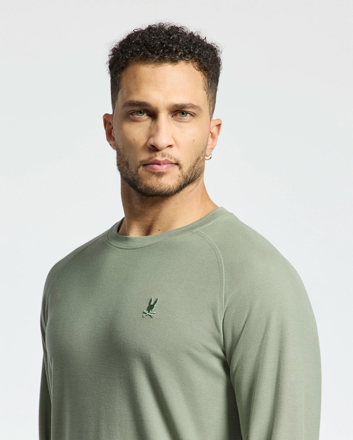 A man with short curly hair and a beard wears the Psycho Bunny's Men's Taylor Pique Long Sleeve Tee in light green, crafted from soft Pima cotton, featuring a small bird logo on the chest against a plain light gray background.