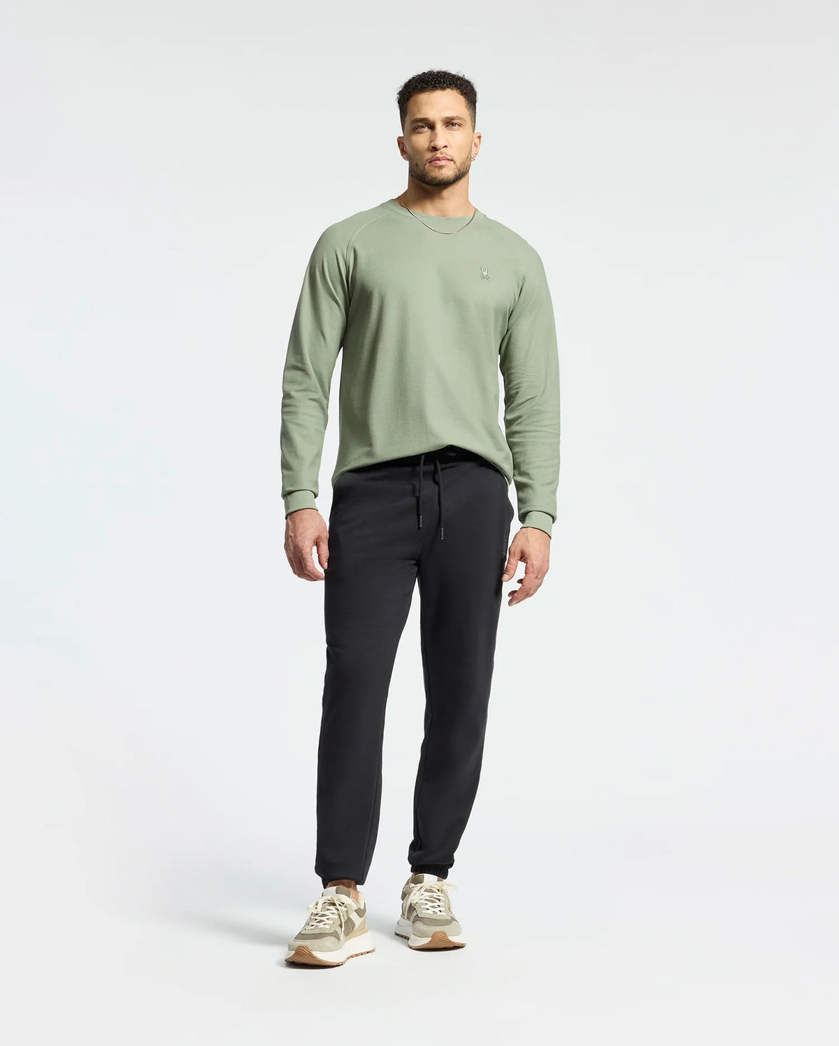 A man stands relaxed in a sage green Psycho Bunny Men's Taylor Pique Long Sleeve Tee, complemented by black joggers and beige sneakers. The plain white background accentuates his casual style.