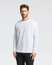 A man with short hair and facial hair wears the Psycho Bunny MENS TAYLOR PIQUE LONG SLEEVE TEE - B6T580F200 in white with raglan sleeves, paired with black pants. He stands against a plain light gray background, slightly facing left, showcasing the embroidered Bunny logo on his chest.
