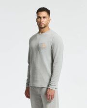 A man stands wearing a stylish Psycho Bunny MENS VAIL WAFFLE LOUNGE LONG SLEEVE TEE paired with matching pants. The light gray shirt, featuring a small orange logo on the chest pocket, adds a touch of flair to his look. A plain, light-colored background highlights the sleek design of this comfortable lounge tee.