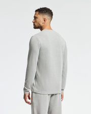 A man with short curly hair stands sideways, wearing a stylish Psycho Bunny MENS VAIL WAFFLE LOUNGE LONG SLEEVE TEE in light gray paired with matching sweatpants. The comfortable ensemble is set against a plain white background.