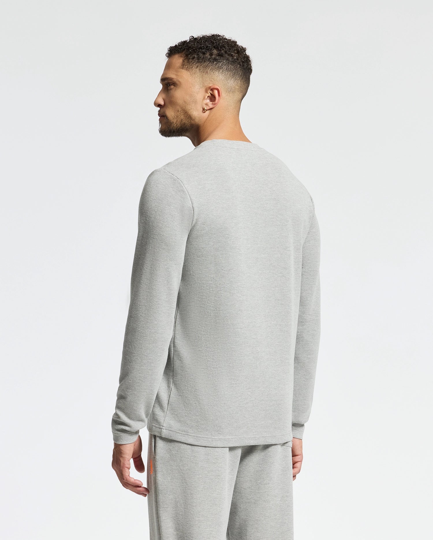 A man with short curly hair stands sideways, wearing a stylish Psycho Bunny MENS VAIL WAFFLE LOUNGE LONG SLEEVE TEE in light gray paired with matching sweatpants. The comfortable ensemble is set against a plain white background.