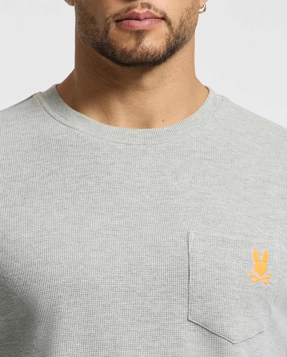 A close-up of a person wearing a stylish Psycho Bunny Men's Vail Waffle Lounge Long Sleeve Tee in light gray. Designed for comfort, the shirt features a small orange rabbit logo above the front pocket. The person's face is partially visible.