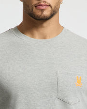 A close-up of a person wearing a stylish Psycho Bunny Men's Vail Waffle Lounge Long Sleeve Tee in light gray. Designed for comfort, the shirt features a small orange rabbit logo above the front pocket. The person's face is partially visible.