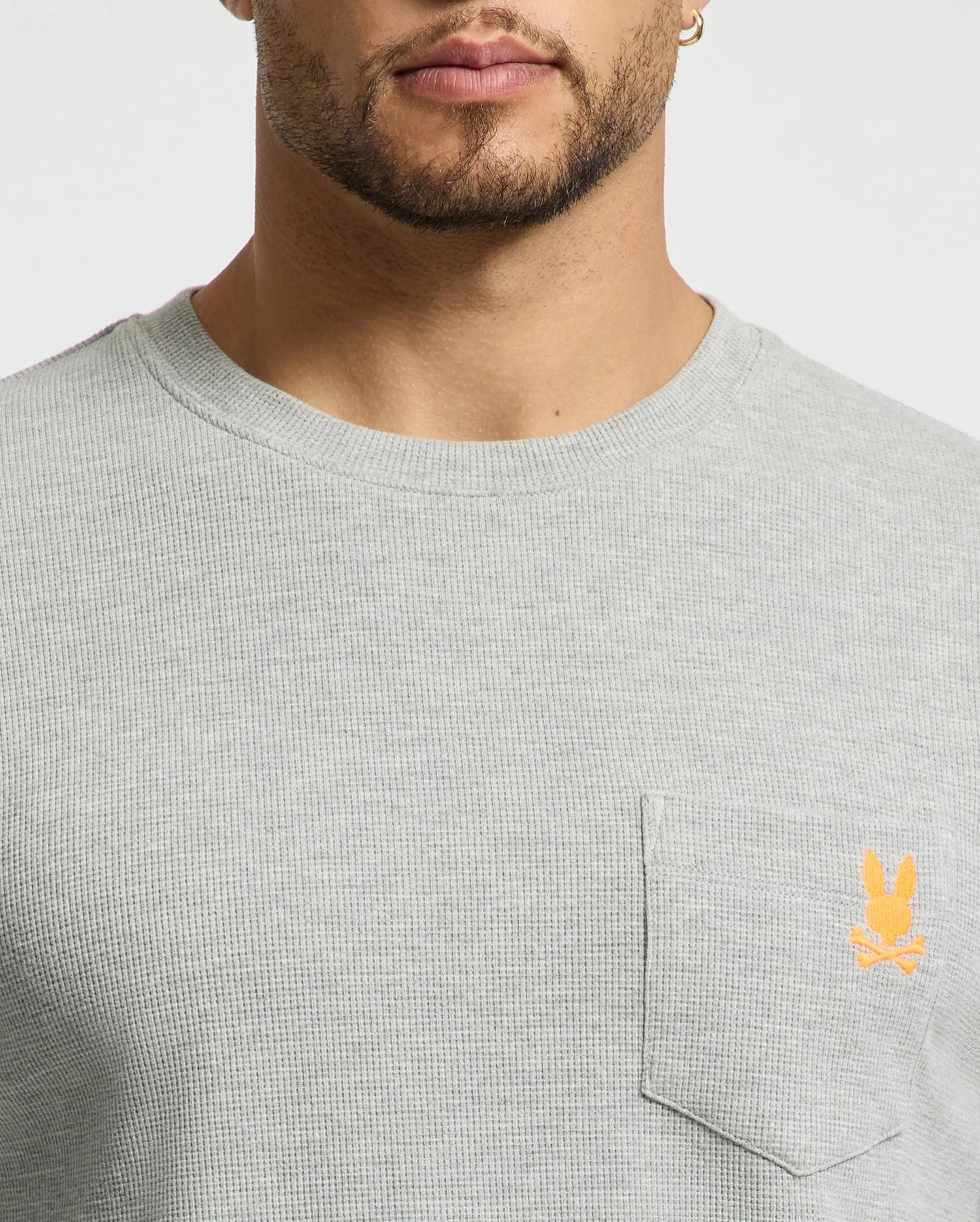A close-up of a person wearing a stylish Psycho Bunny Men's Vail Waffle Lounge Long Sleeve Tee in light gray. Designed for comfort, the shirt features a small orange rabbit logo above the front pocket. The person's face is partially visible.