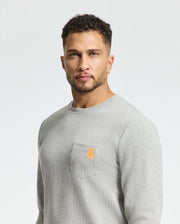 A man with short curly hair and a trimmed beard is wearing a light gray Psycho Bunny Men's Vail Waffle Lounge Long Sleeve Tee, which has a small orange logo on the pocket, set against a plain gray background.