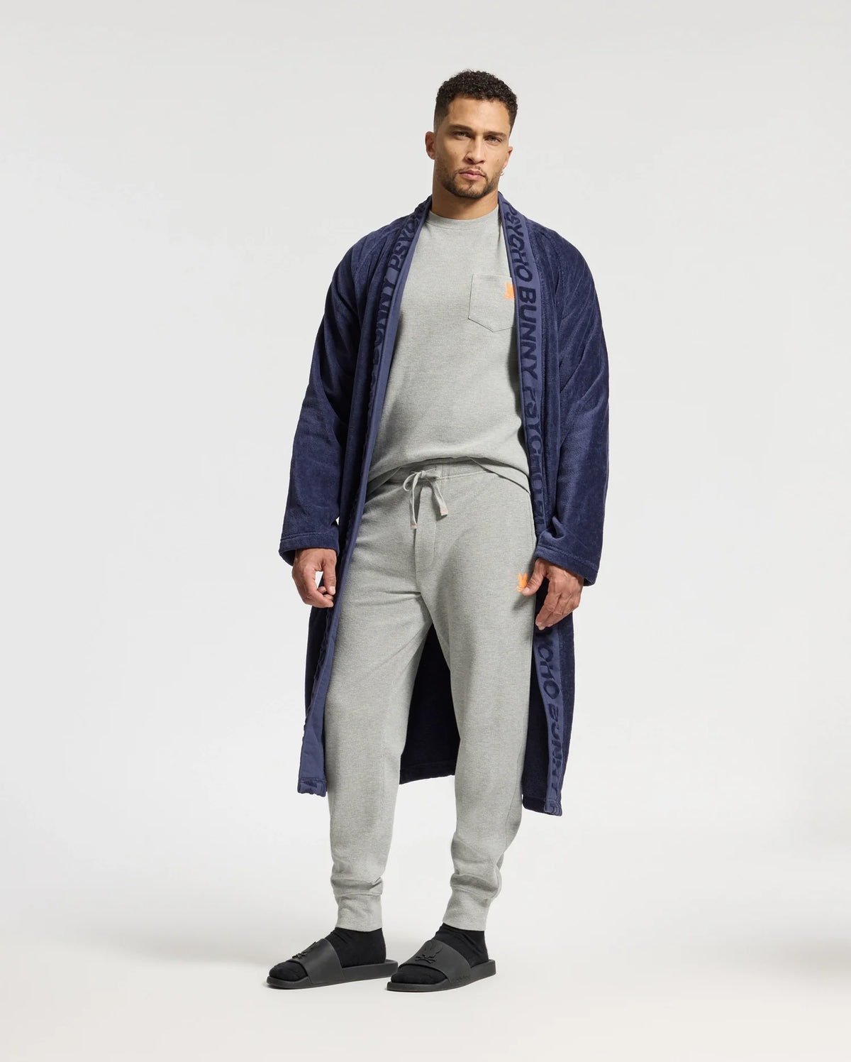 A man stands against a plain background, wearing a navy blue robe with branding on the lapels. Underneath, he is dressed in the Psycho Bunny MENS VAIL WAFFLE LOUNGE LONG SLEEVE TEE – B6T672E200, paired with light gray sweatpants and black sandals, effortlessly blending style with cozy comfort.