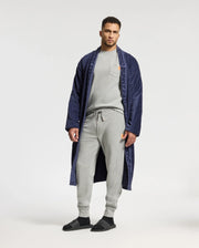 A man stands against a plain background, wearing a navy blue robe with branding on the lapels. Underneath, he is dressed in the Psycho Bunny MENS VAIL WAFFLE LOUNGE LONG SLEEVE TEE – B6T672E200, paired with light gray sweatpants and black sandals, effortlessly blending style with cozy comfort.