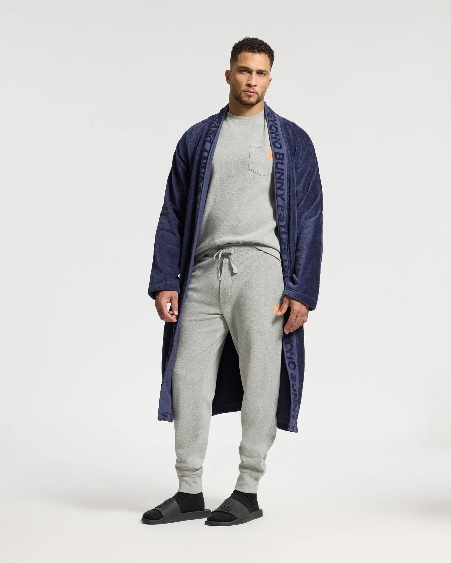 A man stands against a plain background, wearing a navy blue robe with branding on the lapels. Underneath, he is dressed in the Psycho Bunny MENS VAIL WAFFLE LOUNGE LONG SLEEVE TEE – B6T672E200, paired with light gray sweatpants and black sandals, effortlessly blending style with cozy comfort.