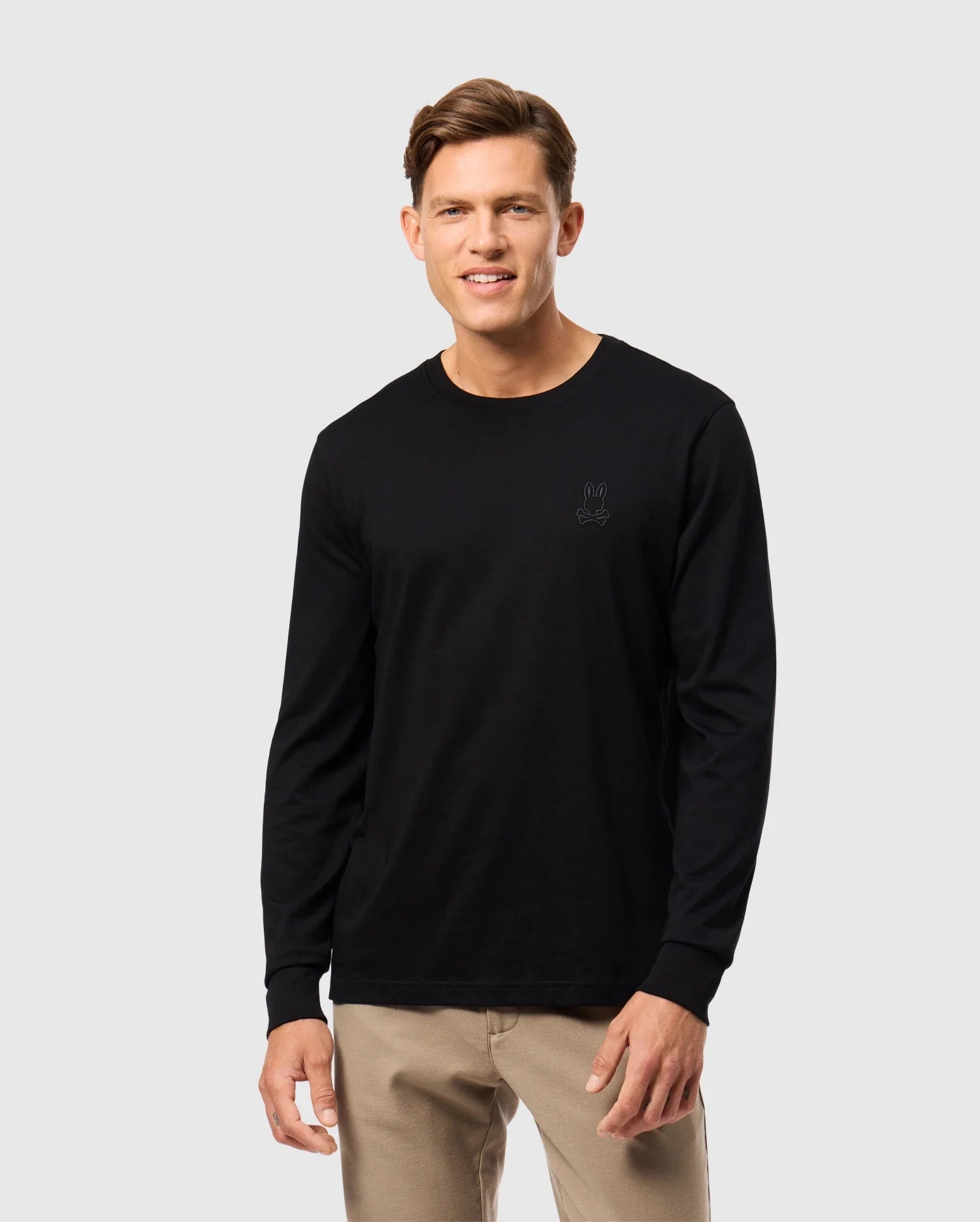 A person stands smiling, wearing a black Psycho Bunny MENS ALBERTON LONG SLEEVE TEE - B6T867D200, crafted from luxurious Pima cotton with a small rabbit logo on the left side, paired with beige pants. The background is plain light gray.