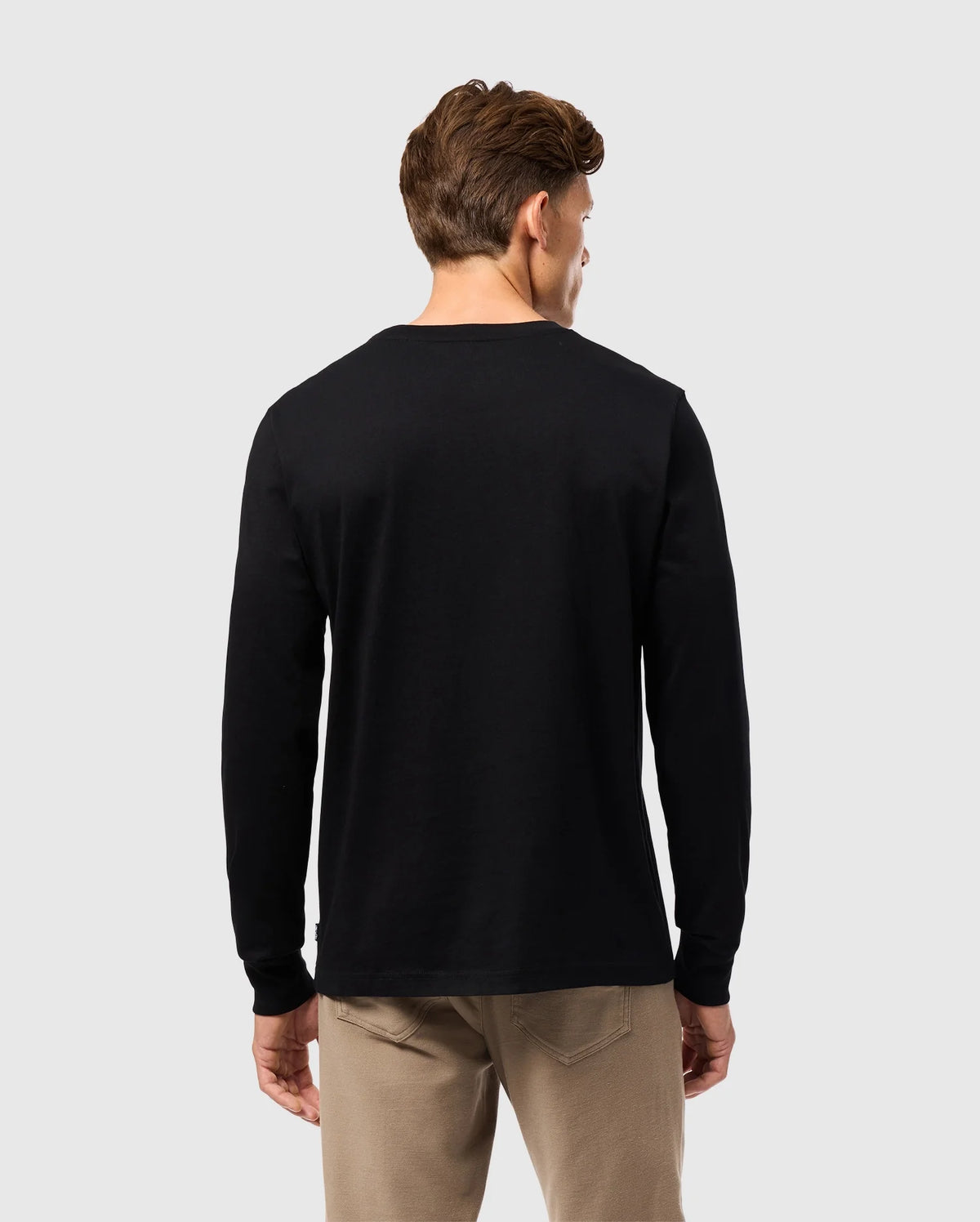 A person with brown hair, wearing a black Psycho Bunny MENS ALBERTON LONG SLEEVE TEE - B6T867D200 and khaki pants, stands facing away against a light gray background.