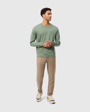 A person wearing the Psycho Bunny MENS ALBERTON LONG SLEEVE TEE - B6T867D200 in green, complemented by beige pants and white sneakers, stands against a light gray background. With short curly hair, they clasp their hands in front, embodying a subtle fashion masterpiece.