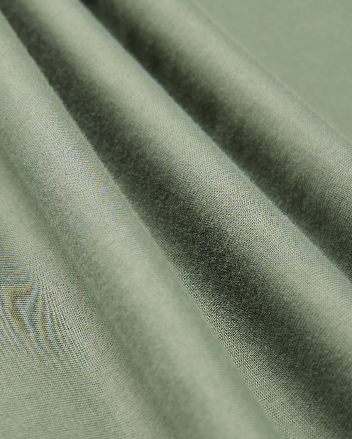 A detailed view of the Mens Alberton Long Sleeve Tee by Psycho Bunny, crafted from green Pima cotton that boasts a smooth texture and soft sheen, beautifully draped in gentle folds. The tee's weave and interaction with natural light produce delicate variations in color and shadow, elevating it to a true fashion masterpiece.