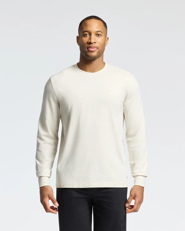 Wearing the Psycho Bunny MENS ALBERTON LONG SLEEVE TEE - B6T867D200 and black pants, a person stands against a light background. This versatile piece complements the outfit as they look directly at the camera.