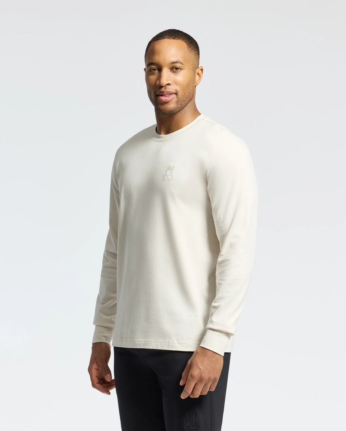 A person wearing a Psycho Bunny MENS ALBERTON LONG SLEEVE TEE - B6T867D200 in white, featuring an embroidered chest design, and black pants stands against a light background. Smiling with one hand in their pocket, it’s the perfect Pima cotton layering piece for casual outings.