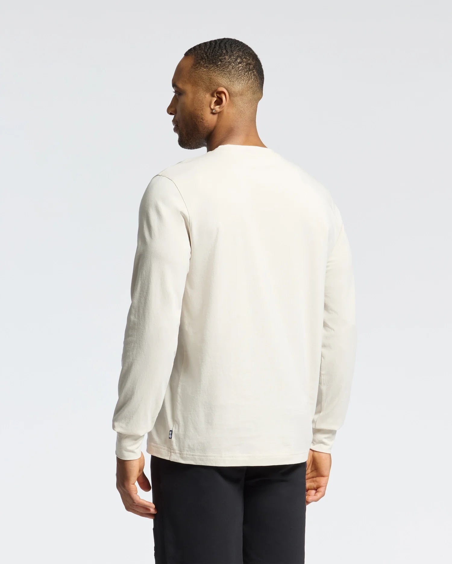 A person in a Psycho Bunny MENS ALBERTON LONG SLEEVE TEE (B6T867D200), white Pima cotton, and black pants stands against a plain background, facing away from the camera, looking to the right.