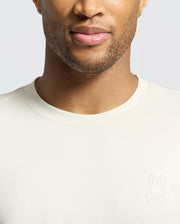 A person wearing a Psycho Bunny MENS ALBERTON LONG SLEEVE TEE in cream, with a bunny and crossbones logo on the left. Made from soft Pima cotton, it highlights the face and upper body against a light gray background, making it an ideal layering piece.