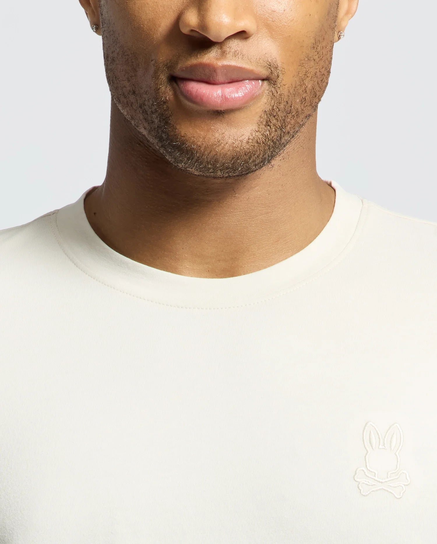 A person wearing a Psycho Bunny MENS ALBERTON LONG SLEEVE TEE in cream, with a bunny and crossbones logo on the left. Made from soft Pima cotton, it highlights the face and upper body against a light gray background, making it an ideal layering piece.