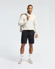 A man in a light sweater and black shorts stands against a white background, holding the MENS ALBERTON LONG SLEEVE TEE from Psycho Bunny over his shoulder, complemented by white socks and sneakers. This classic piece offers comfort and style for any casual outing.