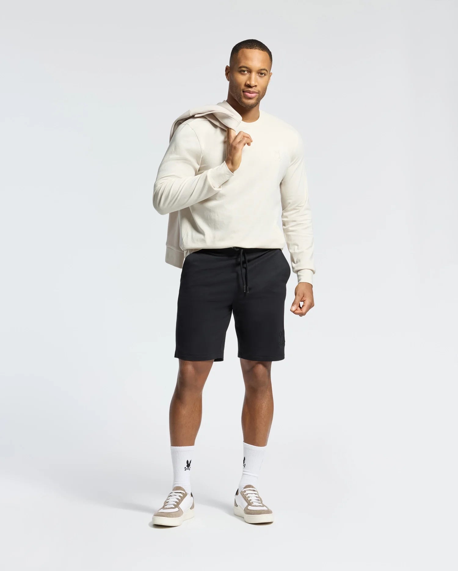 A man in a light sweater and black shorts stands against a white background, holding the MENS ALBERTON LONG SLEEVE TEE from Psycho Bunny over his shoulder, complemented by white socks and sneakers. This classic piece offers comfort and style for any casual outing.