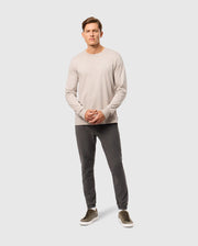 A man leans casually against a plain backdrop, wearing a light gray Psycho Bunny MENS ALBERTON LONG SLEEVE TEE, dark gray pants, and green sneakers. He has short hair and gazes forward with his hands clasped in front.