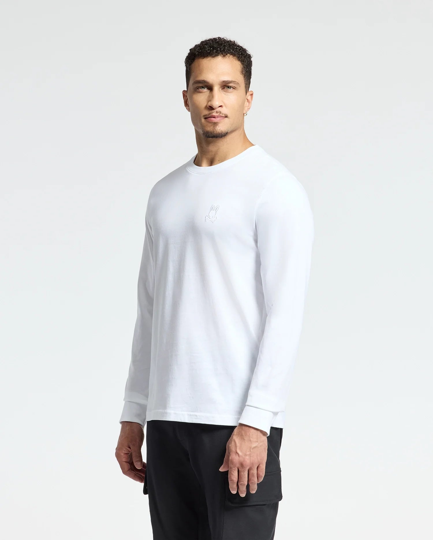 A man with short curly hair is standing against a plain background, wearing the Psycho Bunny MENS ALBERTON LONG SLEEVE TEE in white, featuring a small logo on the chest, paired with black pants. His expression is neutral as he looks directly at the camera.