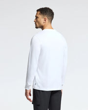 A person with short curly hair is standing slightly turned to the side, wearing a casual Psycho Bunny MENS ALBERTON LONG SLEEVE TEE - B6T867E200 and black pants. The pima cotton shirt adds a touch of elegance against the plain white background.