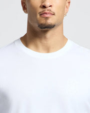 A man wearing a Psycho Bunny MENS ALBERTON LONG SLEEVE TEE in plain white Pima Cotton, highlighted by a subtle bunny logo outline on the chest. Sporting a trimmed beard and a small hoop earring, he stands against a light-colored background.