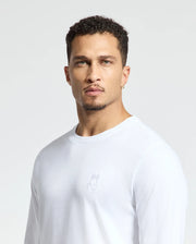 A man with curly hair and a goatee stands against a plain backdrop, wearing the MENS ALBERTON LONG SLEEVE TEE - B6T867E200 by Psycho Bunny. This casual top is crafted from soft Pima cotton and features a small rabbit logo on the chest. He has a neutral expression and wears hoop earrings.