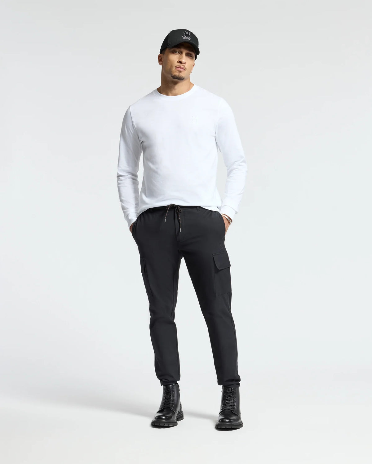 A man stands wearing the Psycho Bunny MENS ALBERTON LONG SLEEVE TEE in white, complemented by a black cap and drawstring black cargo pants. He completes the look with black boots against a plain, light gray background.