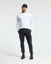 A man stands wearing the Psycho Bunny MENS ALBERTON LONG SLEEVE TEE in white, complemented by a black cap and drawstring black cargo pants. He completes the look with black boots against a plain, light gray background.